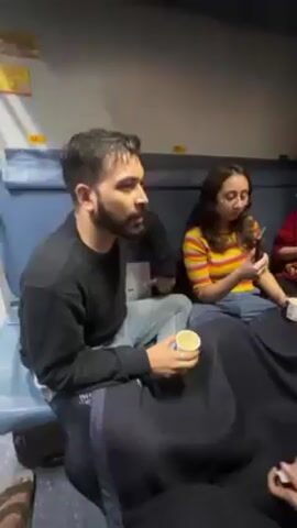Here's a concise description of the video:

The video shows four people sitting in the sleeper compartment of a train. A man on the left wears a black sweatshirt and jeans, holding a cup. A woman in the middle wears a striped shirt and jeans, looking at her phone. Another woman, wearing a red shirt with a cartoon character, sits to the right, also holding a cup. A man on the right is visible at the end, holding cards in his hands. They are all covered by a dark blanket. A small table near the window holds water bottles and other items. The overall environment appears to be cramped and cozy, typical of a train compartment.