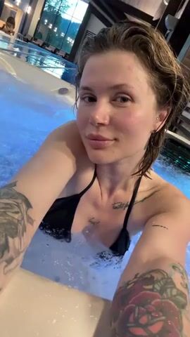 The video shows a woman in a black bikini in a jacuzzi. She is in the foreground, and the background shows an indoor swimming pool area with other people and lounge chairs. The jacuzzi has blue lights at the bottom. The woman is looking at the camera and smiling. She has tattoos on her arms. The video appears to be a selfie-style recording.