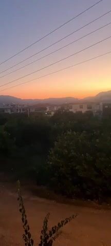 The video shows an outdoor scene during sunset. The sky is a gradient of colors, transitioning from a pale blue at the top to orange and pink closer to the horizon. In the mid-ground, there are trees and some buildings, possibly houses, with lights visible. The foreground is a dirt path with some vegetation. Overhead, power lines are visible against the sky. The camera is static and does not move.