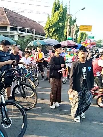 A vertical video shows a dense crowd of people gathered outdoors on a paved road, predominantly on bicycles. In the foreground, a man in a black long-sleeved shirt, a patterned sarong, and a black peci (cap) smiles directly at the camera with his hands clasped together, positioned on a bicycle. To his right, another man in a white t-shirt and blue jeans is also on a bicycle. The crowd, extending into the background, features numerous individuals, many of whom are wearing red and white attire. Some participants are wearing helmets, while others hold umbrellas of various colors for shade. Colorful balloons are also interspersed within the crowd. Trees line both sides of the road, and a yellow road sign is visible in the mid-ground on the left. The event appears to be a large cycling gathering or parade under clear, sunny skies.