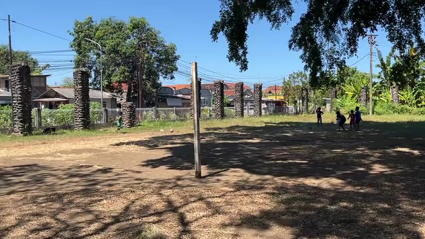 A group of children are playing soccer in an outdoor area. The area appears to be a dirt field with some grass. There is a makeshift soccer net in the foreground, and in the background, there are trees, houses, and a fence. The sky is clear and blue, suggesting a sunny day. The children are wearing casual clothing and appear to be enjoying themselves. One child is actively kicking the ball, while others are positioned around the field. The overall impression is one of a casual, informal game of soccer on a warm day.