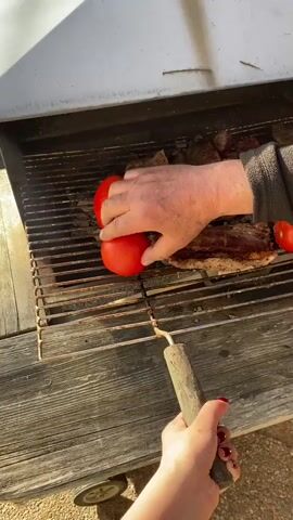 The video shows a close-up view of a grill in Björklinge, Sweden. The grill is filled with several pieces of steak and two red bell peppers and two tomatoes. A person's hands are visible, one placing a bell pepper on the grill and the other holding a long-handled fork, seemingly to rearrange the food. The grill itself appears to be an older model, set on a wooden surface, suggesting it might be outdoors. The lighting suggests it is daytime, possibly late afternoon due to the warm, slightly diffused sunlight. There are no visible signs of text in the media. The overall scene evokes a casual, outdoor cooking experience.