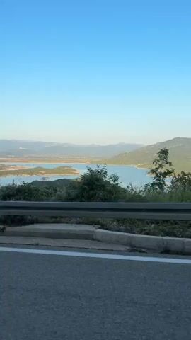 The video is a horizontal, medium-shot, still image of a scenic landscape. The foreground features a gray asphalt road with a white line on the left side and a white concrete curb running along the edge. Behind the road, a metal guardrail separates the road from a grassy embankment. Beyond the embankment, a vast body of water, likely a lake or reservoir, is visible, dotted with small islands covered in green vegetation. The water is surrounded by rolling hills and mountains, with a clear blue sky dominating the upper portion of the frame. A few trees and shrubs are scattered throughout the landscape, adding to the natural beauty of the scene. The image appears to have been captured during daylight hours, likely in the late afternoon or early evening, given the soft lighting and clear sky. No people or specific actions are depicted in the video, suggesting it's a static recording of a natural environment.