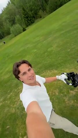 A man is taking a selfie on a golf course. He is wearing a white polo shirt, khaki pants, and white sneakers. He has a golf glove on his left hand. A golf cart with clubs, bags and other accessories is parked on the green. In the distance, other golfers can be seen playing. The sky is cloudy. The setting is a green golf course surrounded by trees.