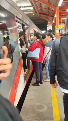 A man in a blue jacket is standing on a train platform, looking at his phone. Several people are boarding a train, including a woman in a red jacket and a woman in a grey top and blue jeans. A train is stopped at the platform, with its doors open. The platform is covered by an orange roof, and there are yellow lines on the ground indicating the edge of the platform.