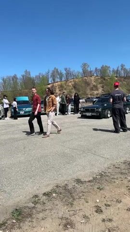 The video shows a group of people standing around several parked cars on a sunny day. The cars are of varying makes and models, including an older, dark green sedan with a classic design. The people are dressed casually and seem to be interacting and chatting. The setting appears to be an outdoor parking lot or gathering space, with a slightly hilly landscape and trees in the background.