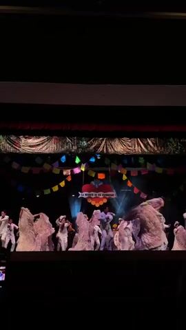 A stage performance shows multiple dancers in what appear to be traditional Mexican outfits. The women wear large, pinkish white dresses, while the men are in white suits. They are twirling and dancing under colorful decorations, including flags and a banner reading "LA MUERTE ES UN FANDANGO" (Death is a Fandango). The stage is illuminated by spotlights, creating a dramatic effect. The scene is filmed from the audience, with a dark foreground showing silhouettes of spectators.