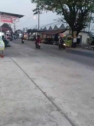 The video depicts a street scene at dusk or dawn. The road is paved with concrete and has painted lines indicating lanes. Several motorcycles are visible, with some riders in motion. A silver minivan drives by on the road. Along the side of the road are various stalls or small businesses, some with awnings and displays of goods. A large tree stands near the roadside, its branches extending over the street. Power lines and poles are present in the background, indicating an urban or semi-urban environment. The sky is overcast or transitioning between day and night. The overall atmosphere is that of a typical street with ongoing activity.
