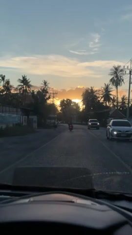 The video shows a view from inside a vehicle looking out at a road during sunset. Palm trees line the sides of the road, and a few buildings are visible. A truck, a motorcycle, and a white car are on the road. The sky is a mix of blue and orange, with scattered clouds. The sun is setting in the distance, casting a warm glow over the scene. The interior of the vehicle is dark, with the dashboard and steering wheel visible in the foreground.
