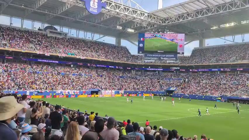 The video shows a wide shot of a crowded soccer stadium during a match. The stands are filled with spectators wearing various colors. On the field, players in white and blue uniforms are visible, engaged in the game. A large screen at the top of the stadium displays the match details, showing a score of 2-0 in favor of the team in white uniforms. The stadium has a modern roof structure and bright lighting. The overall atmosphere appears to be lively and energetic, typical of a professional sporting event.