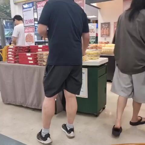 The video captures a casual moment in a retail environment, possibly a bakery or grocery store, in Wonju-si, South Korea. The primary focus is on two individuals, with one person, a man wearing a black polo shirt and grey shorts, holding out two small food items to another person whose hand is visible in the foreground. The man appears to be offering a sample or a treat.

In the background, shelves stocked with various baked goods or packaged foods are visible, suggesting a commercial setting. To the left, a display table is set up with stacked red boxes, and a person in a white shirt is browsing. There is some visible text in Korean on signs behind the display table, including "13,980," which is likely a price. The lighting suggests it is daytime. The overall atmosphere is relaxed and informal, with no indication of a specific event, but rather a typical shopping or browsing experience.
