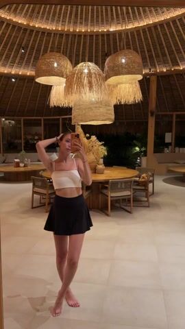 A slender, light-skinned woman is standing in front of a mirror, taking a selfie with her smartphone. She is wearing a white crop top and a short black skirt. Her hair is pulled back. She first has her hand by her head before putting her arms at her sides. The background is an indoor space with a high, straw-covered ceiling and several large, decorative light fixtures hanging from it. There are round wooden tables and chairs arranged in the space, as well as other seating areas. The floor is made of light-colored tiles. Outside of the windows, there is visible greenery and outdoor lighting.