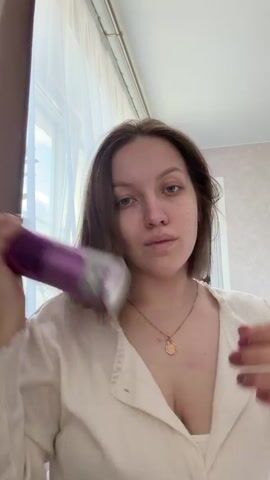 A young woman with shoulder-length brown hair is holding up a purple can of Tigi Bed Head Queen for a Day hairspray and spraying it on her hair. She is wearing a white, V-neck, long-sleeved shirt and a gold necklace with a circular pendant. She has rings on her fingers. She looks directly at the camera as she sprays the hairspray. The background includes a window with white curtains and a patterned wall.