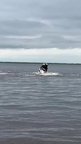 The video captures a person riding a jet ski on a large body of water under a cloudy sky. The jet ski is black with red accents and the rider is wearing a dark outfit with a red helmet. They are creating a significant wake as they move across the water. In the background, a faint green shoreline is visible, suggesting a natural landscape. The overall mood is dynamic and action-oriented, with the weather appearing overcast, which might indicate a cooler or more humid day. The specific location is Ust-Belsk, Russia, though this is not visually evident in the footage itself. There is no visible text in the video.