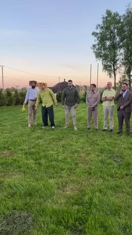The video shows six men on a green lawn under a pale sky. They are all dressed in casual clothing, though some are wearing more formal attire like a suit. From left to right, the first man wears a hat and holds a yellow plastic bag. The second man is in a yellow shirt and dark pants, bending forward. The third man wears a green shirt and beige pants, squatting down. The fourth man wears a light brown shirt and pants and squats. The fifth man in a yellow shirt stands with his arms crossed. The last man is wearing a suit and sits on the ground with his arms open. All the men except the man in the suit are squatting. A few of the men are smiling. Behind the men, there's a small house with a brown roof, trees, and a few houses farther away.