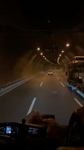 The video shows the interior of a vehicle traveling through a tunnel. The perspective is from the driver's seat, looking forward through the windshield. The tunnel is illuminated by overhead lights, and the walls appear to be made of concrete or rock. There are lane markings on the road. Another vehicle, a red car, is visible ahead in the same lane. A large truck is visible on the right side of the frame, also in the tunnel. The dashboard of the vehicle is partially visible, showing screens and controls. There is a person or a stuffed animal on the dashboard. The lighting is dim, typical of an enclosed tunnel.
