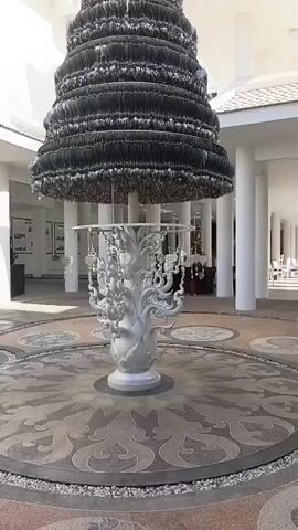 A large, conical sculpture composed of many small, dark elements sits in a circular courtyard. The sculpture's base is a white, ornate pedestal resembling a stylized tree or fountain. The courtyard floor is tiled with a circular mosaic pattern. White, colonnaded buildings with a light-colored roof form the perimeter of the courtyard. Sunlight illuminates the scene. The video is a slow, 360-degree rotation around the sculpture.