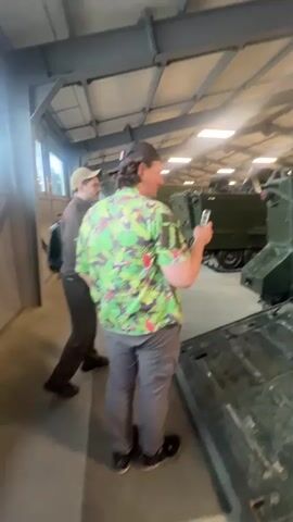 Two men are standing in a large, industrial-looking building with a high ceiling. The man on the left is wearing a dark jacket and a tan baseball cap. The man on the right is wearing a colorful Hawaiian shirt, gray pants, and a dark baseball cap. He is holding a cell phone up and appears to be taking a picture or video of something. In front of them, and slightly to the right, is the open rear of a dark green military vehicle. The interior of the vehicle is visible, painted a light green, with various mechanisms and seating areas. In the background, more of these military vehicles are lined up. The floor is concrete.
