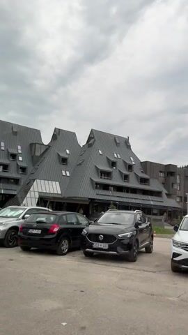 The video begins with a view of a parking lot in front of a uniquely designed building with a grey, angular roof. Several cars are parked in the lot, including a black SUV, a white SUV, and other sedans and hatchbacks. The camera pans to the right, showing more cars and a playground in the background. The building appears to be a hotel or resort, with several stories and many windows. The sky is overcast with clouds. The video then pans back to the left, showing a line of parked cars. The overall scene is one of a well-maintained outdoor area with vehicles and a modern architectural structure.