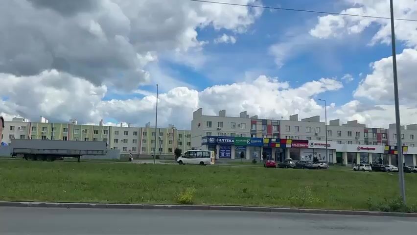 The video shows a long, multi-story building with shops on the ground floor, viewed from across a grassy area and a road. The building has a modern design with repeating sections, some featuring colorful geometric patterns. Several shops are visible, including "АПТЕКА" (pharmacy) and "Пятёрочка" (a grocery store chain). Cars are parked in front of the shops, and a white van is driving on the road in the distance. The sky is blue with fluffy white clouds, and there is a traffic light pole and a transmission tower in the foreground. The video appears to be filmed from a moving vehicle.