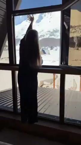 The video shows a young woman standing by a window, looking out at a snowy mountain landscape. She's silhouetted against the bright daylight streaming through the window. She's wearing dark clothing and has long, straight hair.  The window is framed with wood and offers a view of snow-covered buildings nestled in the landscape below the snow-covered mountain. Below the window there is a metal roof visible, and in the background are two hanging wicker chairs. She briefly touches the glass before leaning on the window sill.