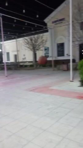 The video shows a nighttime view of an outdoor plaza or park area. The ground is paved with light-colored square tiles, some of which are outlined in red. String lights hang overhead, creating a festive atmosphere.  A claw machine, illuminated with pink and white lights, stands on the edge of the paved area. In the background, there are trees, bushes, street lights, and glimpses of buildings with lighted signs.  A Ferris wheel can be seen in the distance. The scene appears still and quiet.