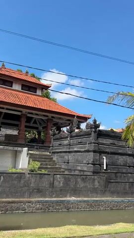 The video pans from left to right across a scene with a clear blue sky and scattered clouds. In the foreground, a paved path runs alongside a narrow canal filled with water. Beyond the canal is a grassy area. A stone wall, likely part of a building, runs along the canal. The wall is dark and intricately carved with decorative elements at the top. A set of stone stairs leads up to an open structure with a red tiled roof. Several palm trees are visible, with one tall tree with green fronds positioned to the right of center. In the background, more buildings with red tiled roofs can be seen, along with lush green foliage. Overhead, power lines are strung across the sky. The overall impression is of a sunny day in a tropical or subtropical location, possibly a temple or historical site. There are no people visible in the video.
