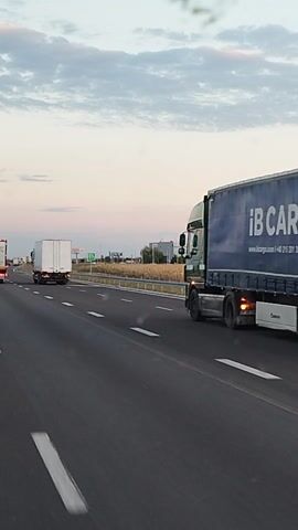The video shows a highway scene in Oarja, Romania. It is likely daytime, given the brightness of the sky and the presence of traffic. The weather appears to be clear with some clouds. The primary subjects are various trucks traveling on the highway. One large green truck is in the foreground on the right, followed by a white truck and then another white truck further down the road. There are also smaller vehicles visible in the distance. The highway has multiple lanes and is marked with white dashed lines. A metal guardrail separates the road from a grassy area with lampposts. In the background, there are some buildings and trees, suggesting an industrial or semi-rural setting. The overall activity is traffic movement on a highway. There is no visible text in the frame. The camera appears to be mounted on a moving vehicle, likely another truck, giving a first-person perspective of the road ahead.