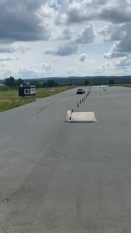 A black car with a number 666 on its rear window performs a drift maneuver on a paved track. The car is moving from the left side of the frame towards the right. Orange cones are lined up along the side of the track. A white barrier is positioned on the track in front of the car. In the background, there are fields, hills, and a cloudy sky. A building with a large tent-like structure is visible in the far distance. The track appears to be an outdoor course, possibly a racetrack or a testing ground.