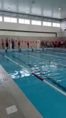The video shows an indoor swimming pool with lane markers dividing the pool.  A group of people, some dressed in swimming attire, are gathered at the far end of the pool near the starting blocks. Some appear to be coaches or spectators. There is a lifeguard standing near the side. A row of triangular flags decorates the wall behind them.  Sunlight filters through large windows. The camera pans slightly across the scene.