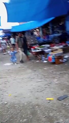 A busy outdoor market scene is depicted. The ground is dirt and littered with trash. A blue tarp stretches overhead, providing shade for stalls. In the foreground, a young child in a blue dress is being led by the hand by an adult male in a striped shirt and light-colored pants. They are walking towards the left of the frame. Several market stalls are visible, displaying produce, boxes, and other goods. People are browsing and working at the stalls.