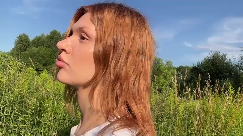 A close-up shot focuses on a young woman with long, reddish-blonde hair walking through a grassy field under a bright blue sky. The sun illuminates her face, highlighting her skin and hair. She turns her head slightly, looking towards the camera with full lips and clear eyes. Tall green grass and foliage surround her, with trees visible in the background. The lighting suggests it is daytime, likely late afternoon or early morning due to the warm glow on her face.