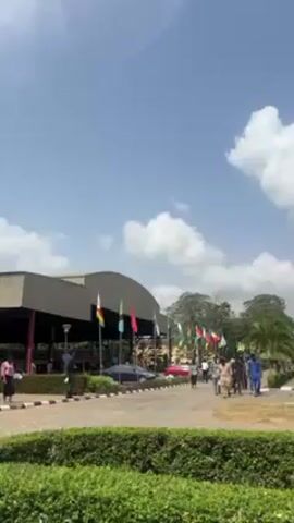 The video shows a building decorated with flags and people walking. The building is a large, low structure with a curved roof. There are several flags of different nations or organizations displayed in front of the building. People are walking along the road in front of the building. The sky is blue with white clouds. The scene is likely a public event or gathering. There's a well-maintained hedge in the foreground. The weather is sunny. A multi-story building can be seen to the right, with people standing outside. A bus is also visible in the background.