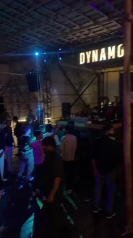 The video shows a lively scene inside a club or bar. The ceiling is constructed from what appears to be bamboo or thatched material. A large illuminated sign reading "DYNAMO" is visible at the back of the venue, above a DJ booth where several people are gathered. A DJ is actively mixing music, as indicated by the equipment on the booth. A crowd of people, mostly young adults, are dancing and socializing. Blue and green laser lights are projected across the room, illuminating the dancers and creating a dynamic atmosphere. Some individuals are holding drinks. The overall impression is that of a bustling nightlife venue with music and dancing.