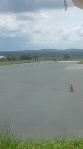 A dark-colored car drives on a paved surface, navigating a course marked by orange traffic cones. The car is traveling from left to right in the frame, appearing to make a turn as it progresses. The environment is outdoors under a cloudy sky, with a grassy foreground and a treed, hilly landscape in the background. A few small structures are visible in the distance. The car appears to be alone on the course.