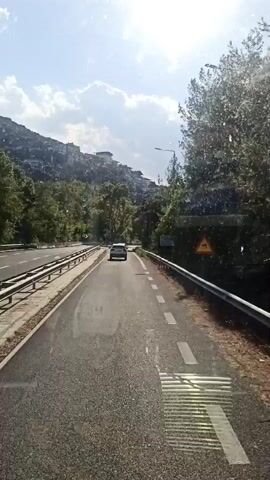 The video shows a view from inside a vehicle traveling on a road. The road is paved and has white lane markings. There are guardrails on both sides of the road. On the left side of the road, there is a hillside covered with trees and buildings. On the right side of the road, there are trees. In the distance, there are two white cars driving on the road. The sky is blue with some clouds. The sun is shining.