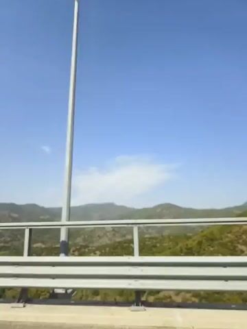 The video shows a view from a moving vehicle on a bridge. In the foreground, there is a metal guardrail. Beyond the guardrail, there are green trees and rolling hills. The sky is clear blue with a few scattered clouds. A tall pole is visible on the right side of the frame. The camera appears to be moving forward.