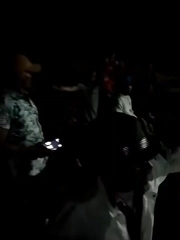 This is a video of a night-time event. The first few seconds show a group of people standing in the dark, some holding cell phones. The scene then transitions to a concert or festival. A stage with bright lights is visible in the background, with a band or performers on it. A large crowd of people is gathered in front of the stage, with many of them silhouetted against the lights. A large screen is also visible on the left side of the stage, displaying an image. The video appears to be filmed by someone in the crowd, as the camera is moving and shaking. The lighting is dim, with the stage lights being the primary source of illumination. The ground appears to be uneven and possibly muddy.