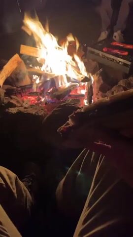 The video shows a campfire at night. Logs and embers glow red and orange as flames leap upwards. There are rocks around the fire pit. In the foreground, a person's hands are visible holding food, possibly a hot dog, over the fire. To the right, a grill with several hot dogs cooking over the fire is visible. In the background, a person wearing a dark jacket and light-colored pants stands and appears to be holding a flashlight. Another person is partially visible to the right. A white vehicle with its headlights on is also visible in the background. The overall environment appears to be a campsite or outdoor gathering at night.