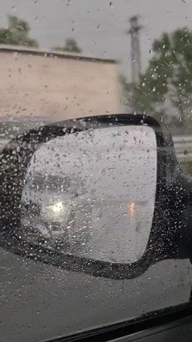 The video shows a view from inside a car during a rainstorm. The camera is looking out of the side window and into the side mirror. Water droplets cover the glass and the mirror. The side mirror reflects the road ahead, showing other vehicles and the rainy environment. The video also shows a silver SUV driving on the wet road, kicking up spray from the puddles. The trees in the background are blurred by the rain and the motion of the car. The overall impression is one of driving in heavy rain.