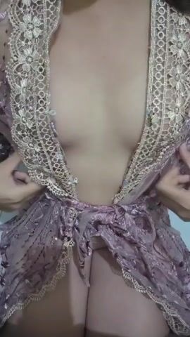 A person wearing a sheer, purple, lacy robe with gold trim is shown from the chest down. The robe is open, revealing cleavage and the top of the person's legs. The person's hands are adjusting the robe. The background is a plain, light-colored wall.