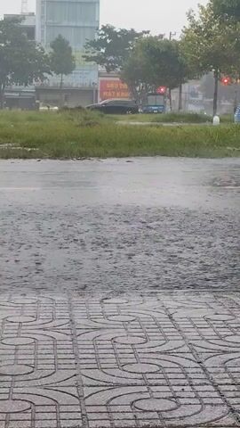 This is a vertical, medium shot of a street during a rainstorm. The foreground is a wet, patterned sidewalk. Beyond the sidewalk is a wet road with visible raindrops hitting the surface. In the middle ground, a person on a scooter rides by, wearing a light-colored rain jacket and helmet. The scooter is dark-colored. In the background, there is a grassy area, trees, and buildings. There are also cars and traffic lights visible. The overall impression is of a heavily raining day in an urban or suburban setting.