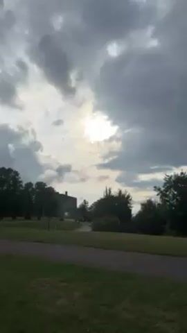 The video shows a partly cloudy sky above a grassy hill, with a paved path cutting across the foreground. Behind the hill, several buildings are visible, including one with a prominent steeple, situated in a city. Trees are scattered throughout the scene. The sun is obscured by the clouds, but its position can be inferred from the brighter areas in the sky.