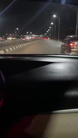 The video shows a car driving at night. The view is from inside the car, looking out the windshield. The dashboard is black, and the exterior is lit up by streetlights. Other cars are on the road, with their headlights and taillights visible. The road has a median strip.