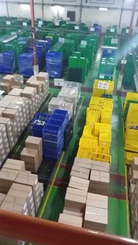 The video shows a high-angle view of a large warehouse or distribution center. The floor is a bright green with yellow and red lines indicating pathways or zones. Rows of neatly stacked cardboard boxes are visible in the foreground. Beyond the boxes, there are stacks of blue and yellow plastic bins, some of which are filled with items. In the background, numerous rows of large, green bins or containers stretch into the distance. The overall impression is one of organized storage and inventory management. There are no people visible in the video. The lighting appears to be artificial, consistent with an indoor industrial setting.