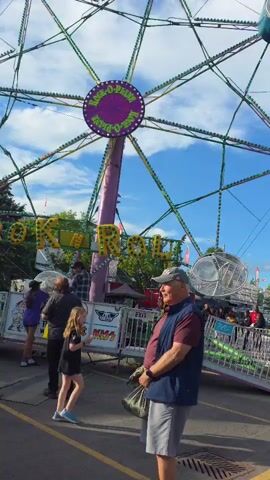 A group of people stand near a carnival ride with a sign that reads "LOCO-MOTION". The ride is a large Ferris wheel-like structure with gondolas. A man in a blue vest and grey shorts is in the foreground, holding a bag. A young girl in a black dress walks past him. The sky is blue with some clouds. There are other carnival rides and tents in the background.