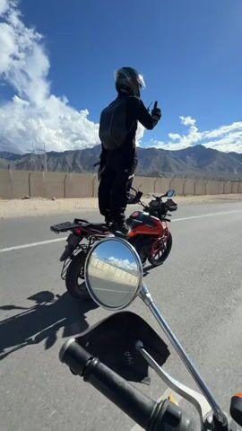 A person on a motorcycle is standing on the seat of the motorcycle, wearing a helmet and backpack, and giving a thumbs up. The motorcycle is parked on the side of a road. In the foreground, a motorcycle mirror is visible. In the background, there are mountains and a blue sky with clouds. The video appears to be taken from another motorcycle moving alongside the parked one.