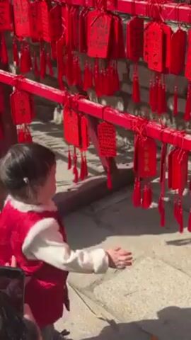 A small child in a red vest is standing in front of a red railing covered in red tags with tassels. The child is reaching out to touch one of the tags. The railing is made of metal and the tags are made of paper or cardstock. The ground is paved with stone. Another adult can be seen in the very lower left corner of the frame, possibly filming the child. The background appears to be a building with stone walls. The child appears to be exploring the tags. The scene is bathed in bright sunlight.