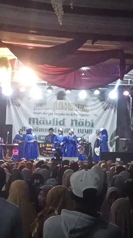 The video shows a stage performance in front of a large crowd. On the stage, there are several people dressed in blue outfits, some holding microphones and musical instruments. There is a large banner behind them with text that appears to be in Arabic and Indonesian. The crowd in front of the stage is packed, with many people wearing headscarves. The lighting suggests it is nighttime or an indoor event. The overall atmosphere appears to be a religious gathering or celebration.