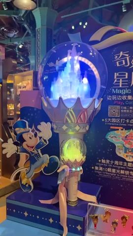 A video shows a display of Disney merchandise, possibly in a store. In the foreground, a Mickey Mouse figure in a blue and gold magician's outfit stands on a blue platform with gold stars. To the right of the Mickey Mouse figure is a large, clear orb on a stand. Inside the orb is a miniature castle illuminated with white and blue lights. Below the orb is a smaller wand with a yellow light. The background features a large sign with Chinese characters and English text that reads "Magic star wand" and "Play, Collect, Connect." The sign also includes images of Disney attractions and characters. Below the main display, a shelf holds small figurines of Disney characters. The lighting in the scene is dim and atmospheric, with the lights from the display being the primary light source. The overall impression is of a themed retail environment.